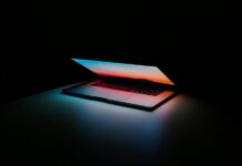 Nuove tecnologie e progresso: cosa cambierà nei prossimi anni Open silver laptop on a dark desk, screen showing a sunset gradient and blue/orange glow on the surface; the room is mostly dark.