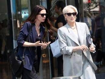 Two stylish women walking outside a storefront; the one on the left wears a dark blue blouse and sunglasses, the one on the right wears a light gray blazer and sunglasses.