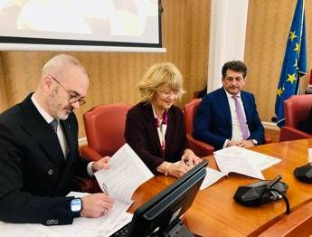 Three people sit at a wooden conference table signing documents; a laptop and papers are visible, with an EU flag in the background.