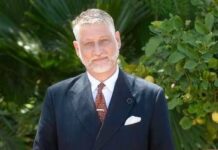 Cultura, Giuli: "Sterilizzati tagli per luoghi memoria, confidiamo in legge bilancio" Man in a dark suit, white shirt, and brown tie stands outdoors against lush green foliage.